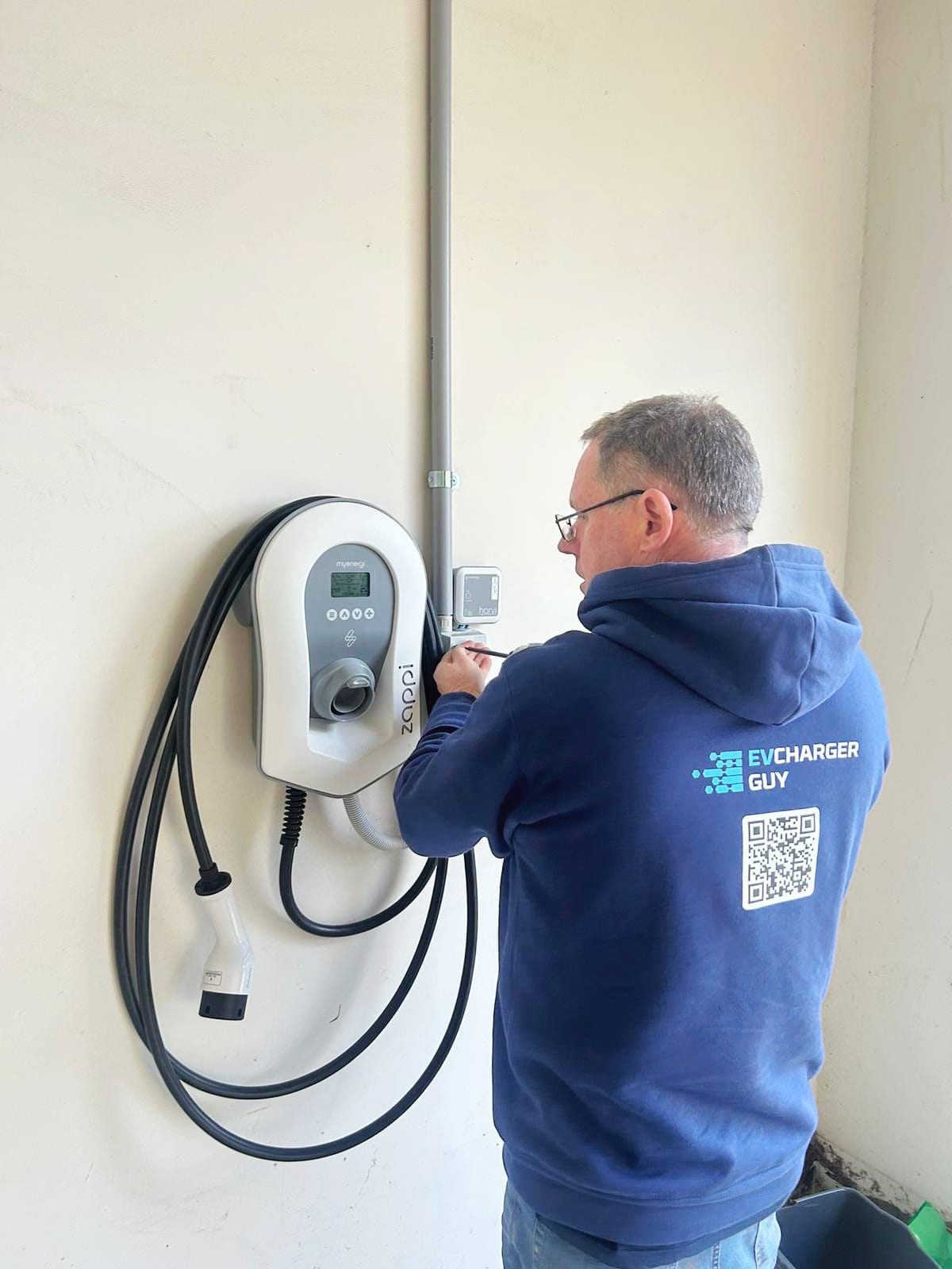 Person in a blue hoodie with 'EV Charger Guy' branding working on an electric vehicle charging station.